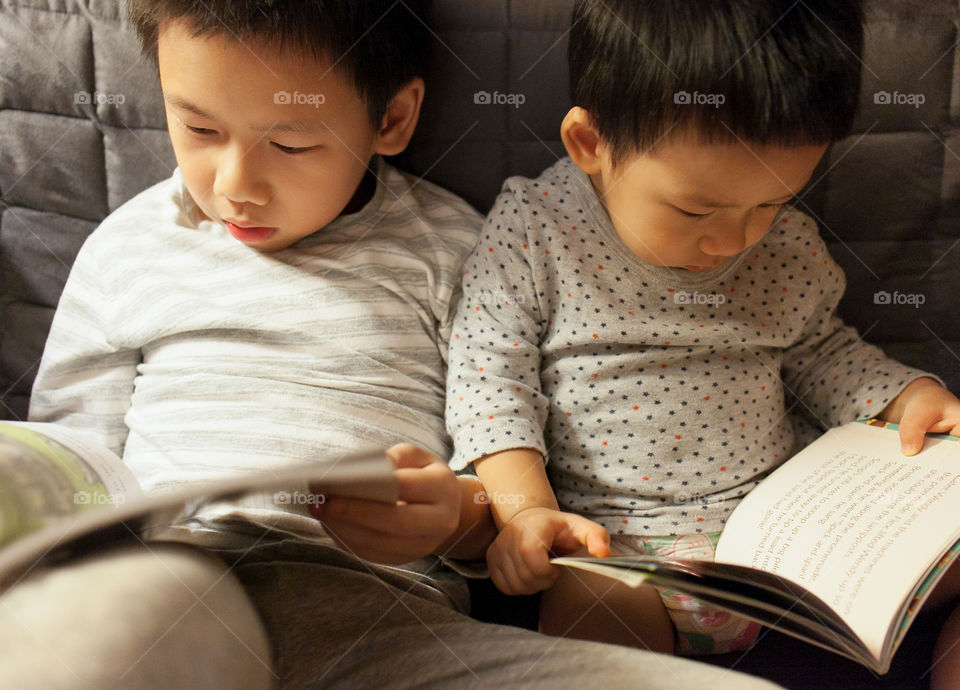 Adorable brothers in pyjamas reading books before bed on the couch, side by side.