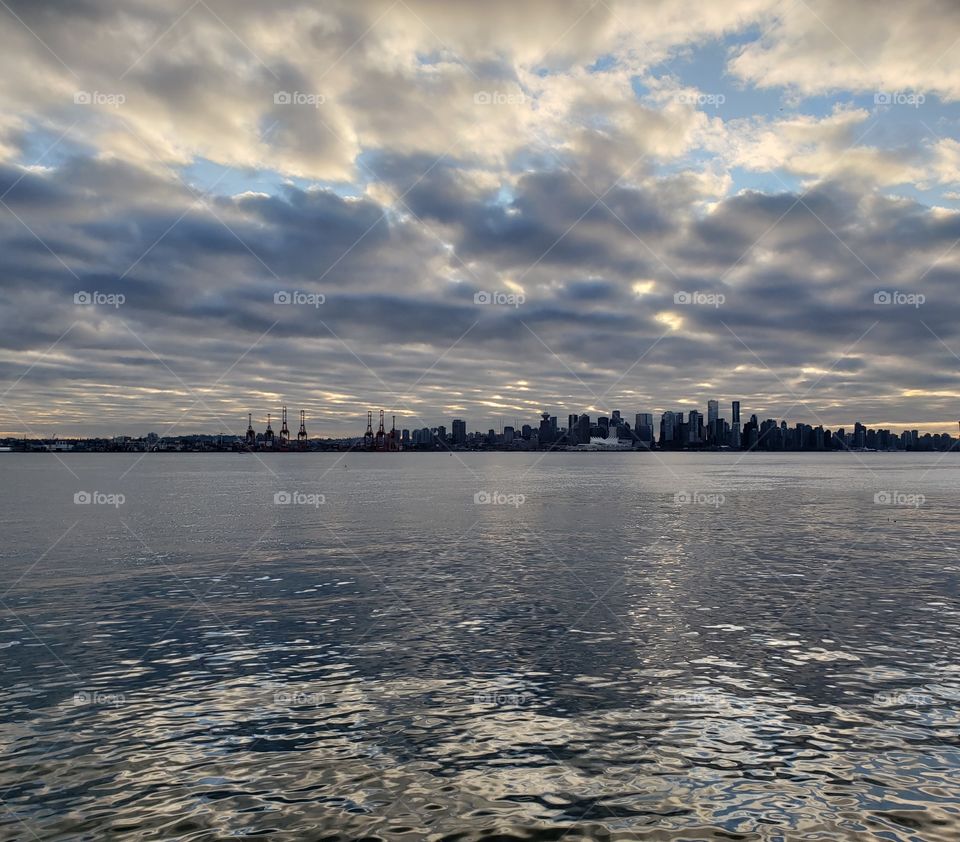 unique magical sky, sun glows through clouds over reflections in wavy ocean water and silhouette of city landscape, shades of Blue, gray, and white