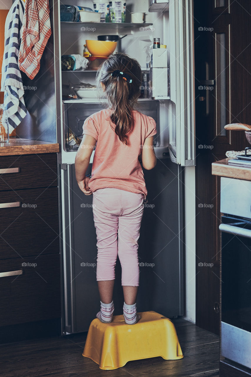 Little girl standing on a childs stool in front of opened fridge, looking for ice creams among food products. Real people, authentic situations