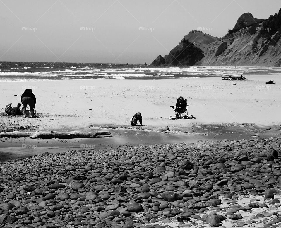 People playing and relaxing on a beach filled with sand and rocks with a stream flowing through and surrounded by cliffs and the Pacific Ocean of the Oregon coast on a sunny summer day. 
