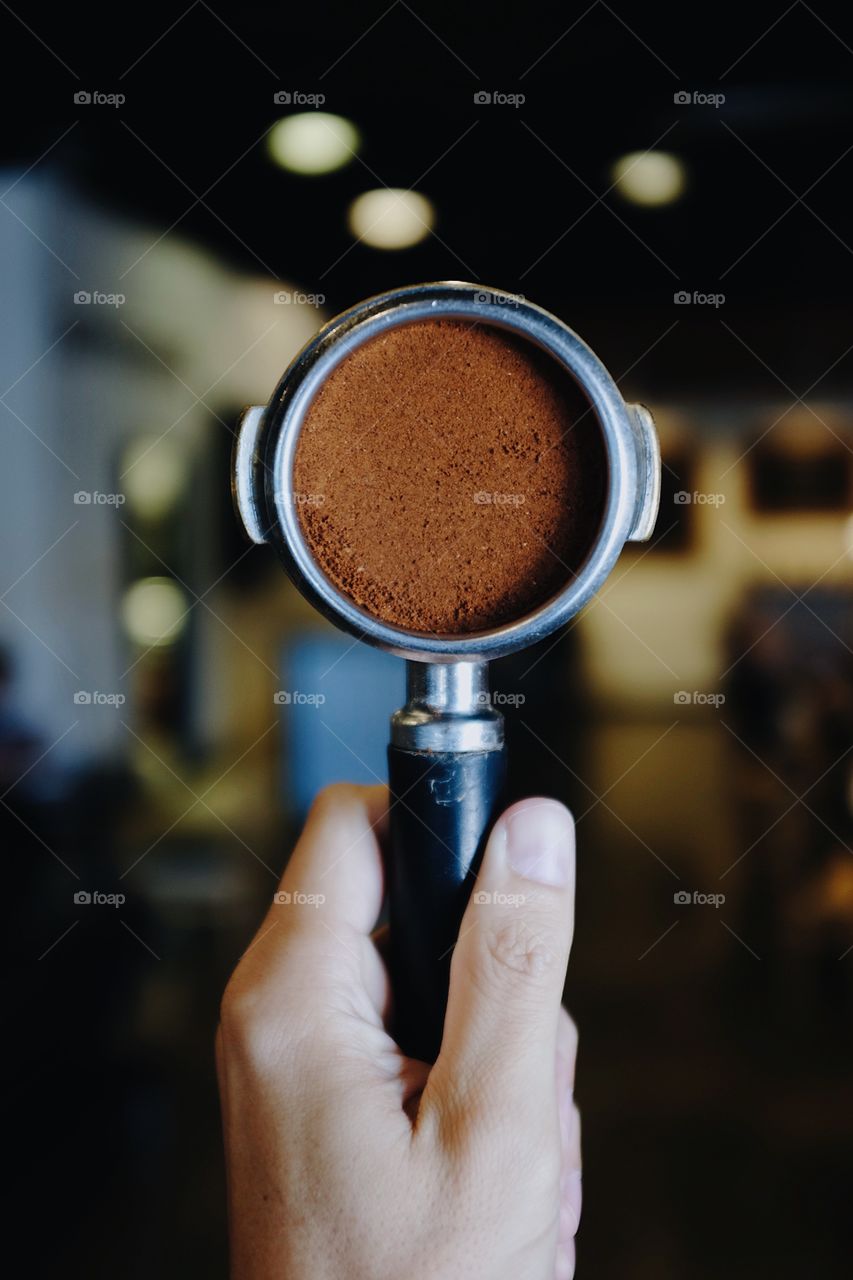 Hand holding a portafilter filled with espresso coffee with a bokeh background inside of a coffee shop. 