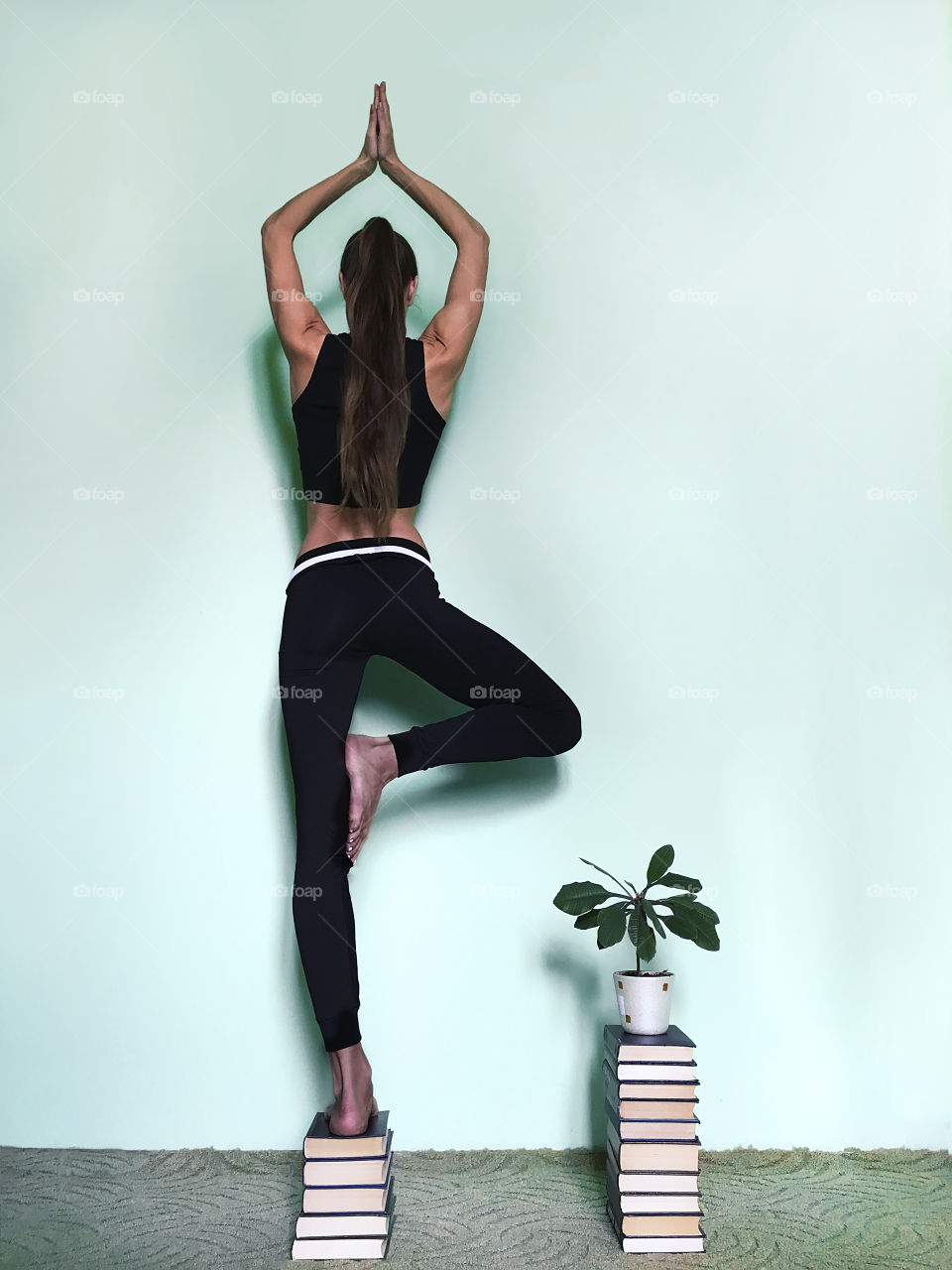 Young woman with long hair doing yoga next to the house plant in front of the blue monochrome wall 
