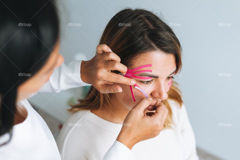 Young beautiful brunette woman with long hair doctor cosmetologist makes face taping for patient in office. Young women do anti-aging procedures with help of kinesio tape