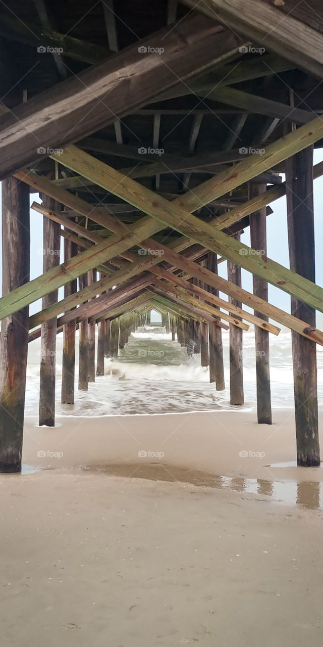 waves crashing against the posts underneath a pier on the beach.