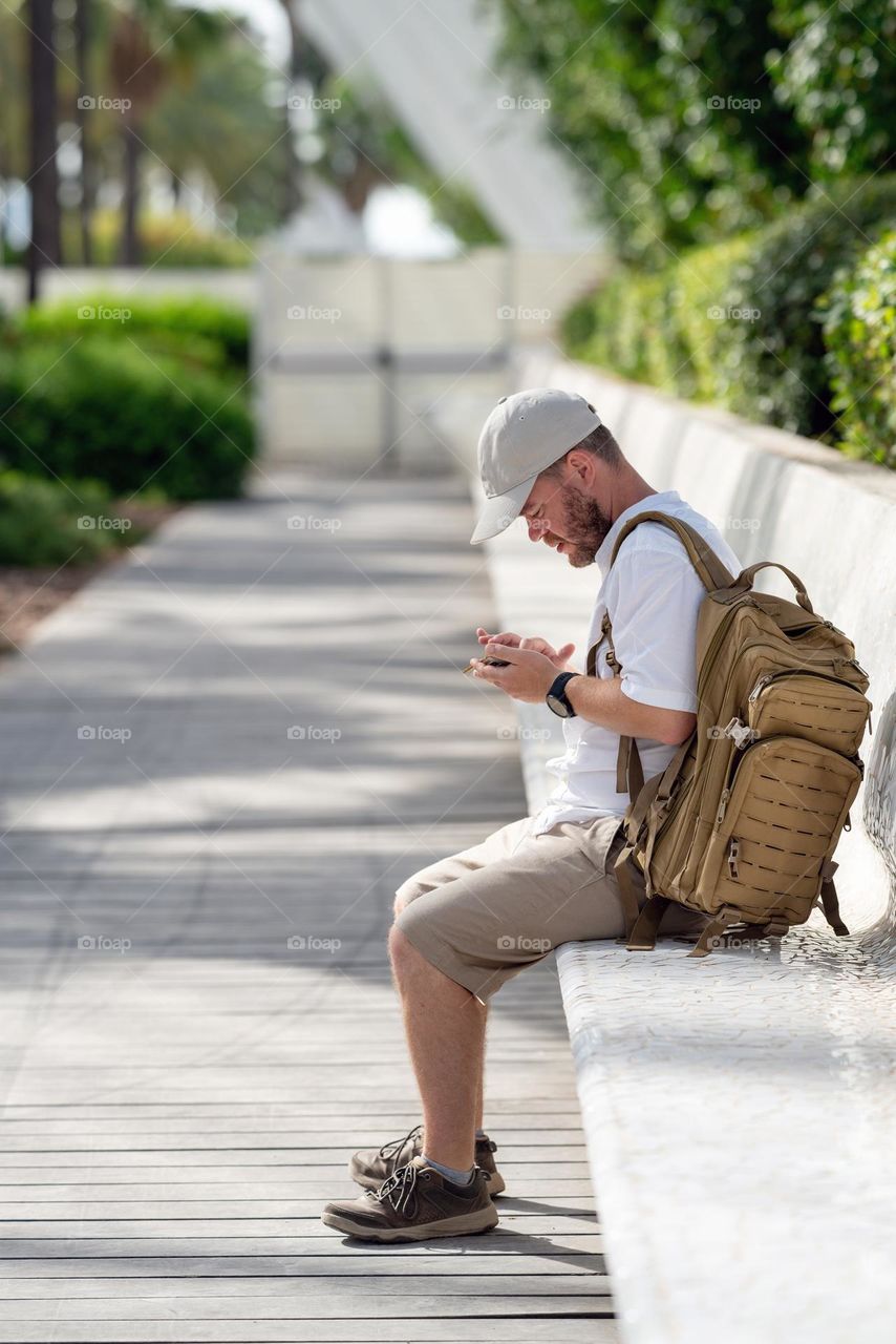 Man in casual clothes using smartphone. Tourist with backpack sitting in the park.