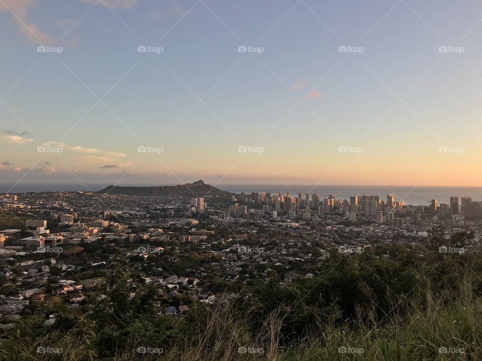A cityscape of downtown Honolulu at sunset. Diamond Head can be seen overlooking the ocean in the distance.