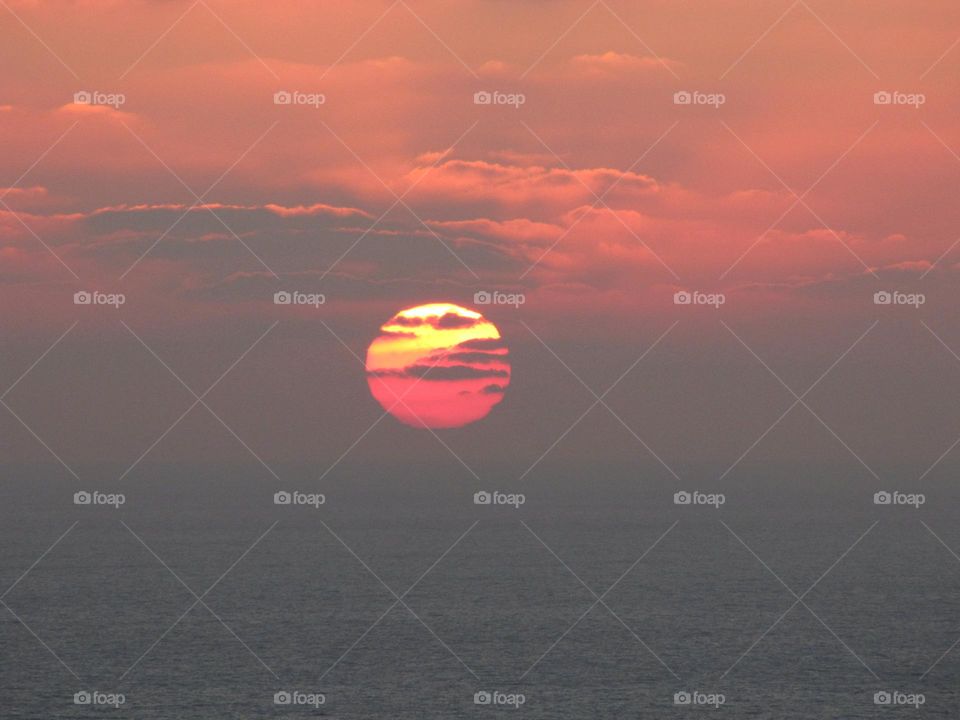 The red sun at sunset (it seems to be on fire) over the sea highlighting and coloring the sky and the clouds around it. Amazing pink, red, yellow and orange.