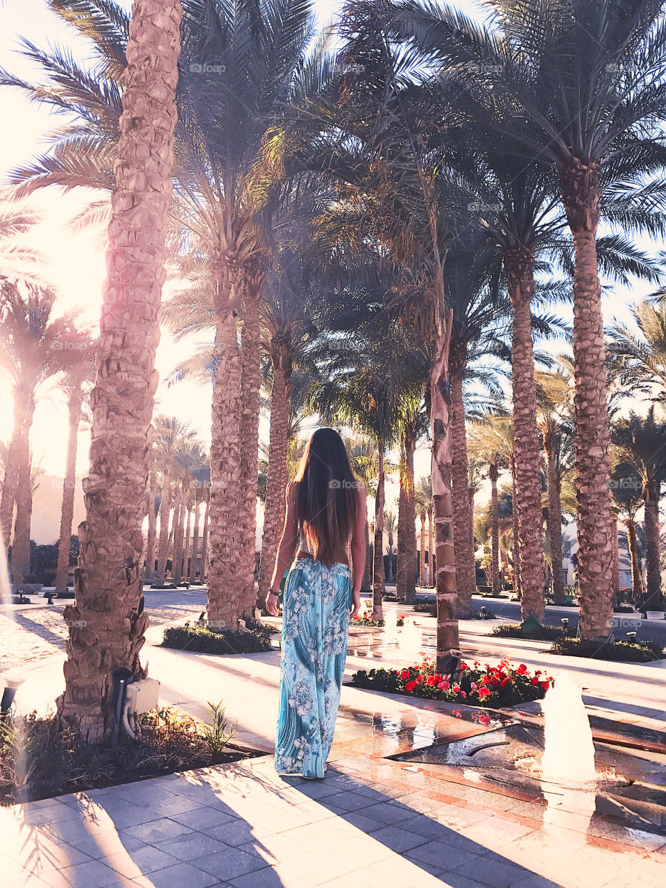 Young woman with long hair wearing a blue dress and walking through the tropical palm trees 
