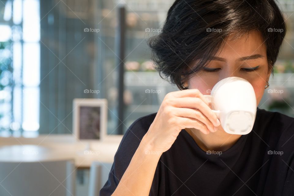 A beautifil asian woman happily sipping coffee in a cafe' with her eyes closed joyfully.
