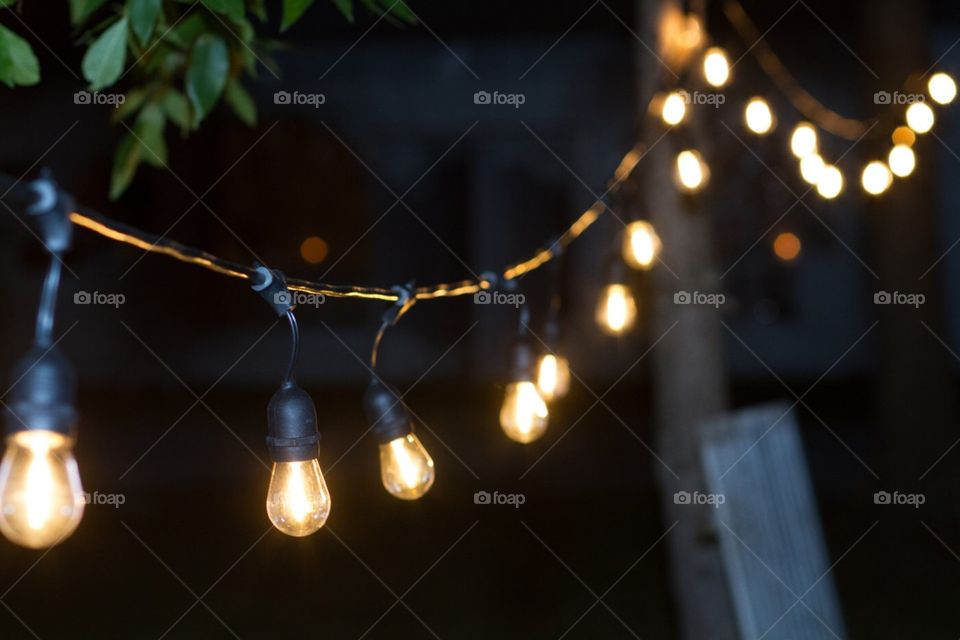 A string of vintage and retro edison lightbulbs lines and lights the path to a wedding tent in the distance at the wedding party reception in the tree lined park