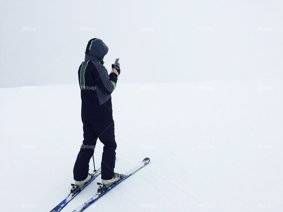 Skier in black ski costume with smartphone in his hand texting surrounded by fog and snow