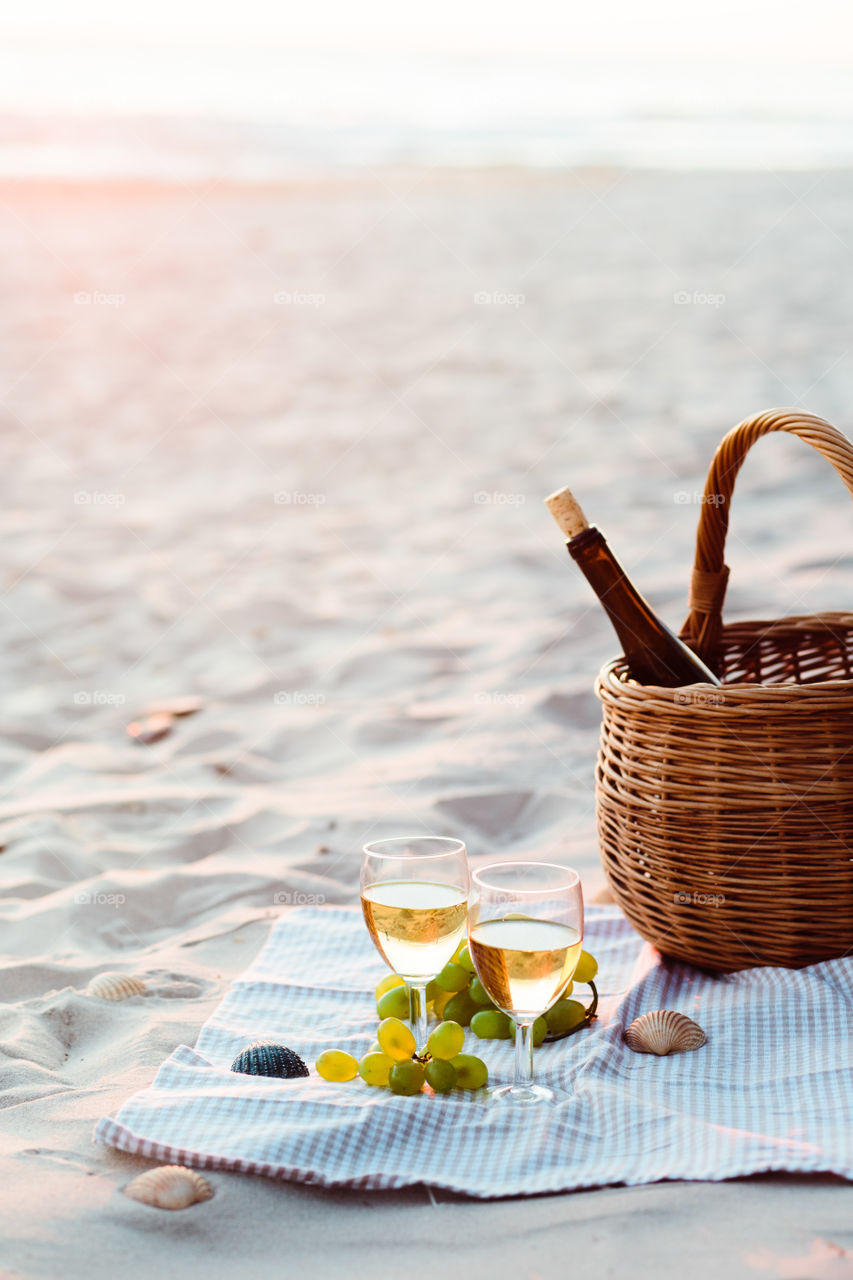 Two wine glasses with white wine standing on sand, on beach, beside grapes and wicker basket with bottle of wine. Sea waves in the background