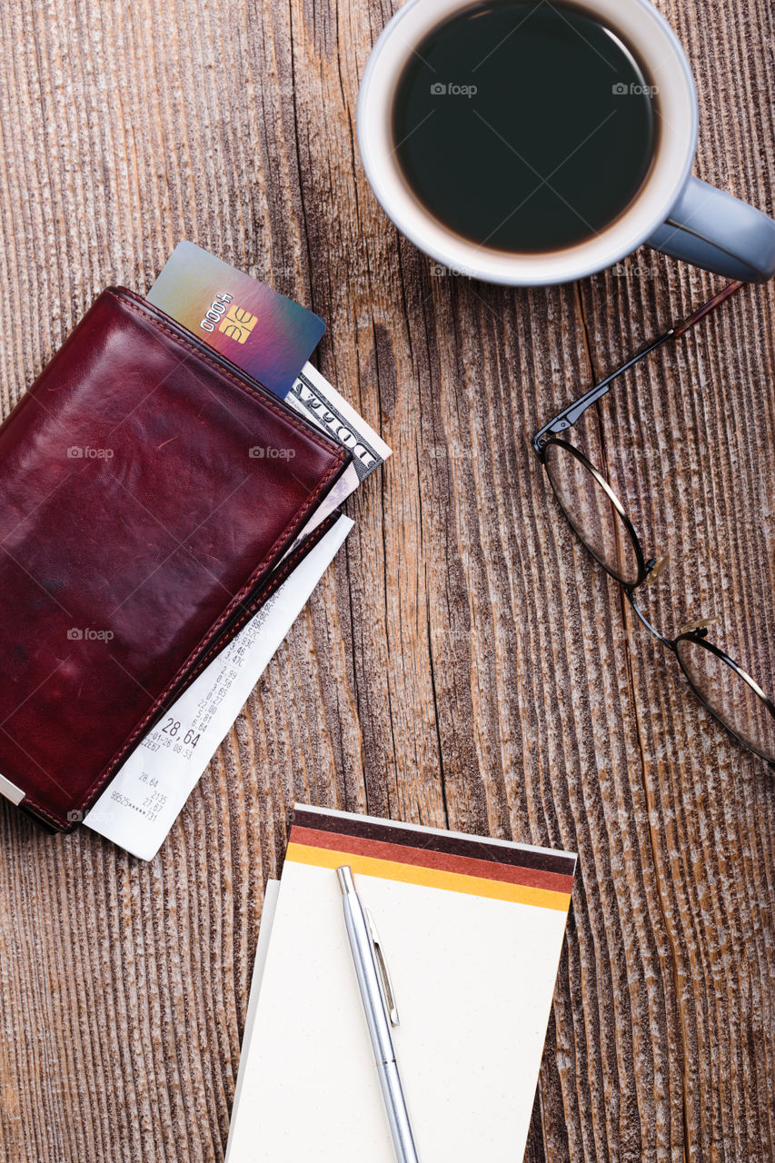 Leather wallet with dollar banknotes, credit card and receipt, glasses, cup of coffee on wooden desk. Portrait orientation