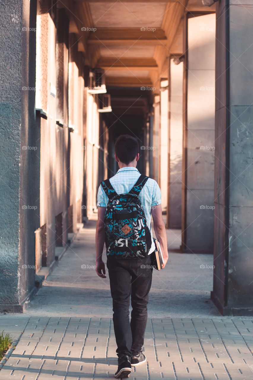 Student holding a notebook and carrying a backpack walking at the front of university building. Young boy wearing blue shirt and dark jeans. Back view