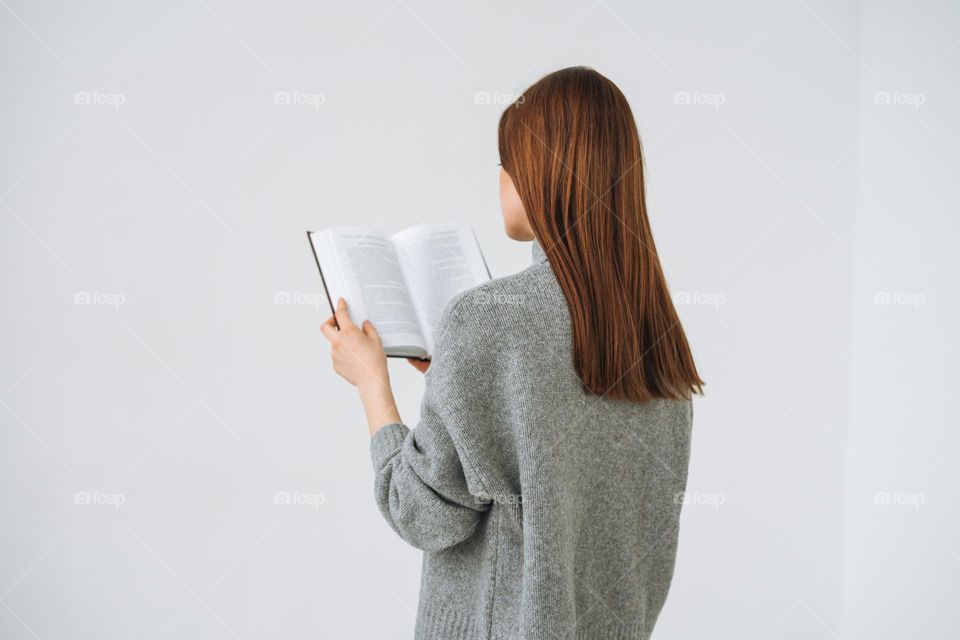 Young woman girl with dark long hair in grey knitted sweater reading book on white background 
