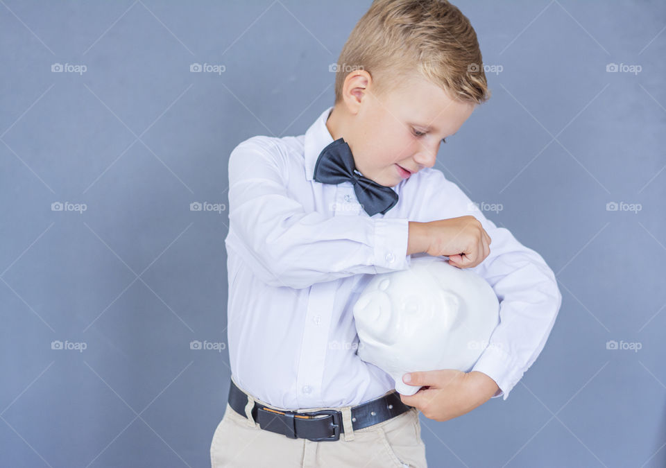 Stylish and positive schoolboy in a white shirt with a saving bow tie and a piggy bank piggy bank coin glass on a blue background for saving money for future use, finance accounting concept copy space  