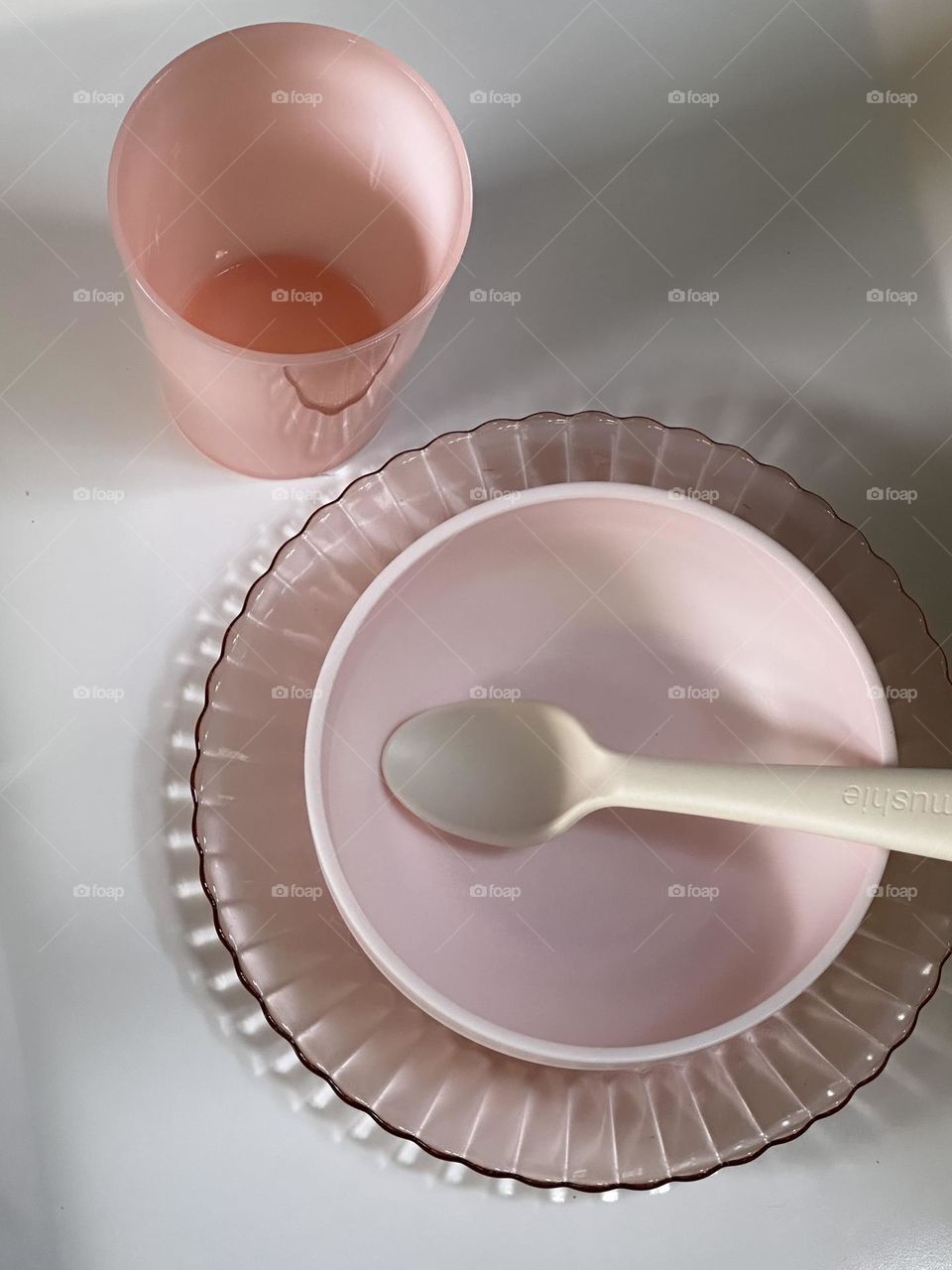 Pink plastic bowl with off white plastic spoon on pink glass plate with peach translucent plastic cup on white background 