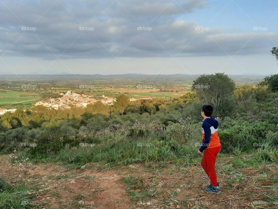 Boy at an elevated position on an hill, looking at a village situated at a lower position. Vegetation around and some clouds at the sky are visible. Santa Eugenia, Majorca (Spain)