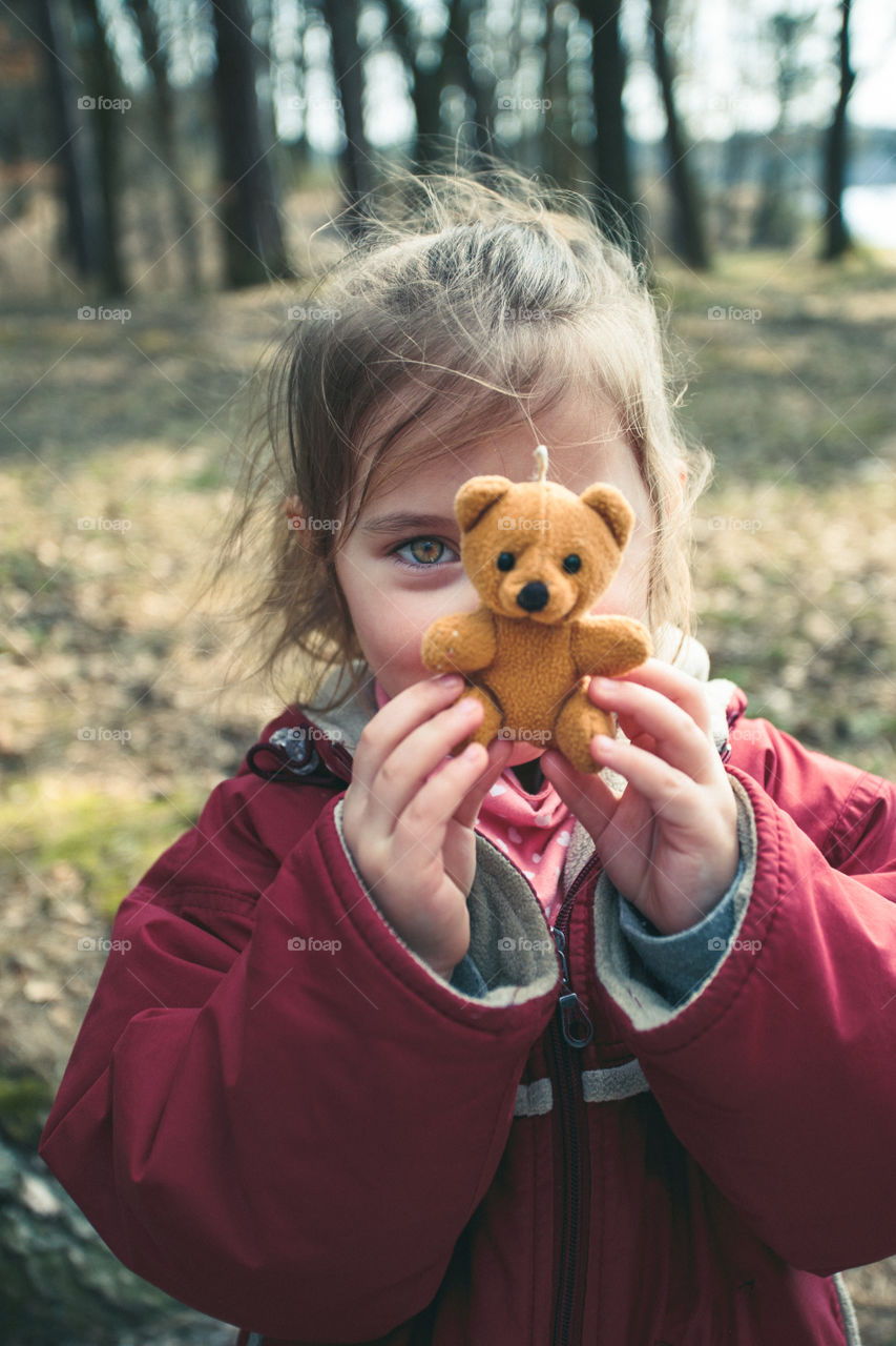 Little smilling happy girl playing with her little teddy bear toy in a park on sunny spring day. Child holding teddy at front of face looking at camera wearing red jacket