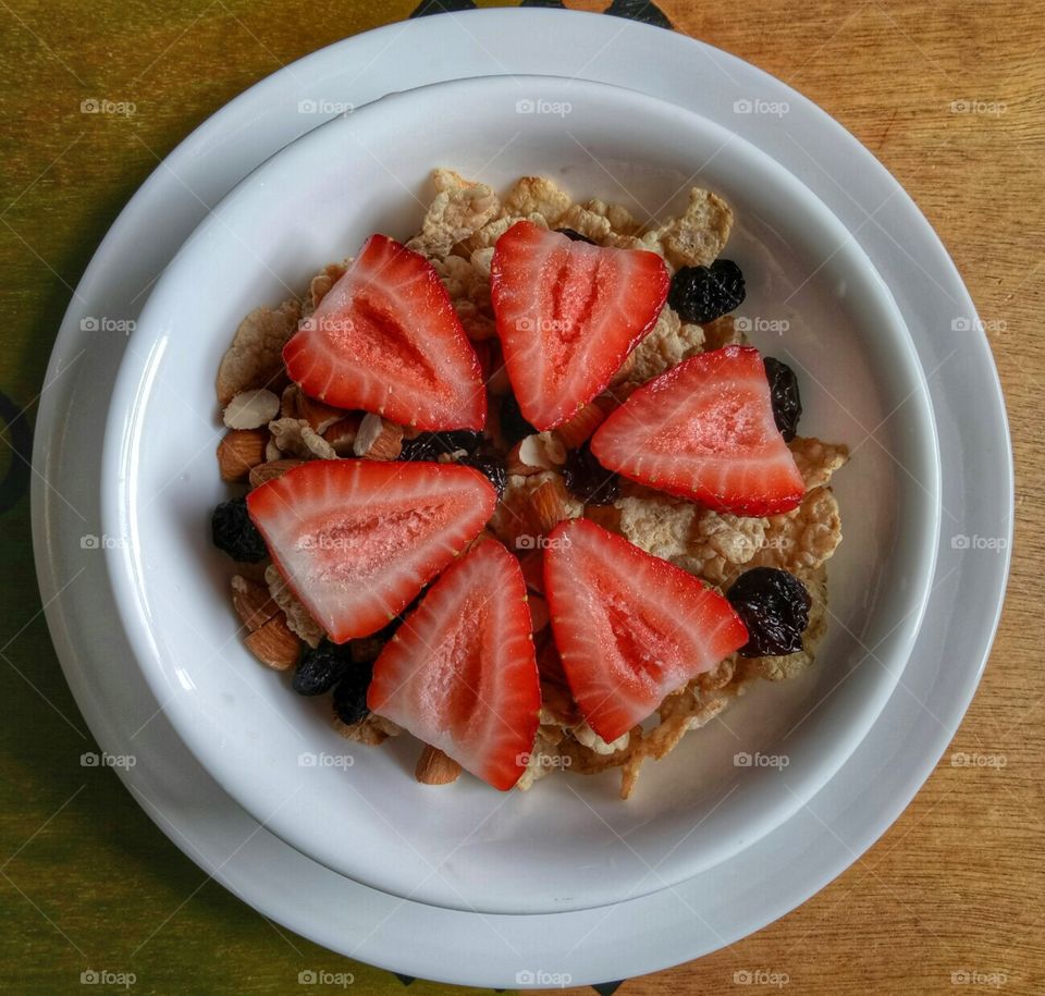 Slice of strawberries, raisins, cereal, almonds in a white round bowl in a brown background.