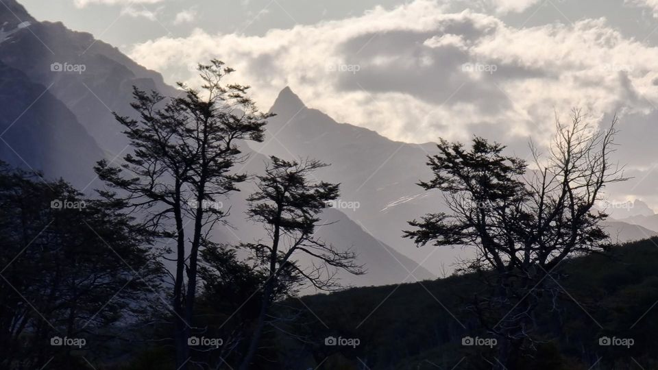 the dark silhouette of some trees and in the background the silhouette of the mountains and some clouds in the sky highlighted by the sun peeking out from behind.