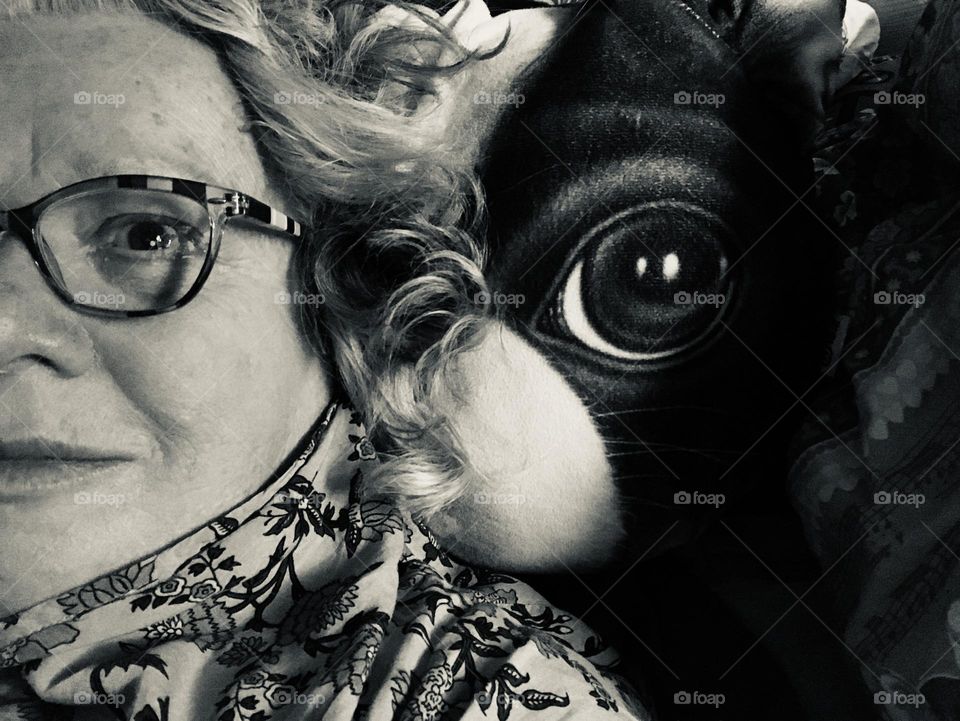 Silver tone selfie of the left side of the face of a woman in glasses lying next to her big eyed Boston Terrier pillow. Her wavy hair creates a soft boundary so that only the left side of the pillow is shown. Double reflections shine in both eyes. 