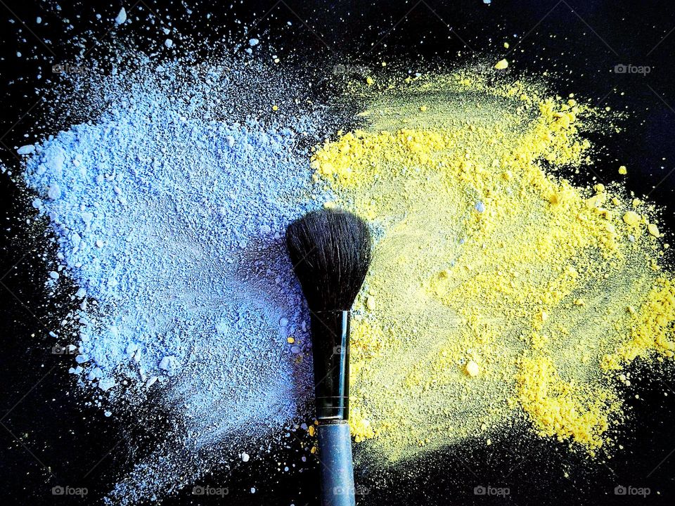 Decorative cosmetics. Blue and yellow shadows are scattered on a black background. There is a black tassel on the border of colors