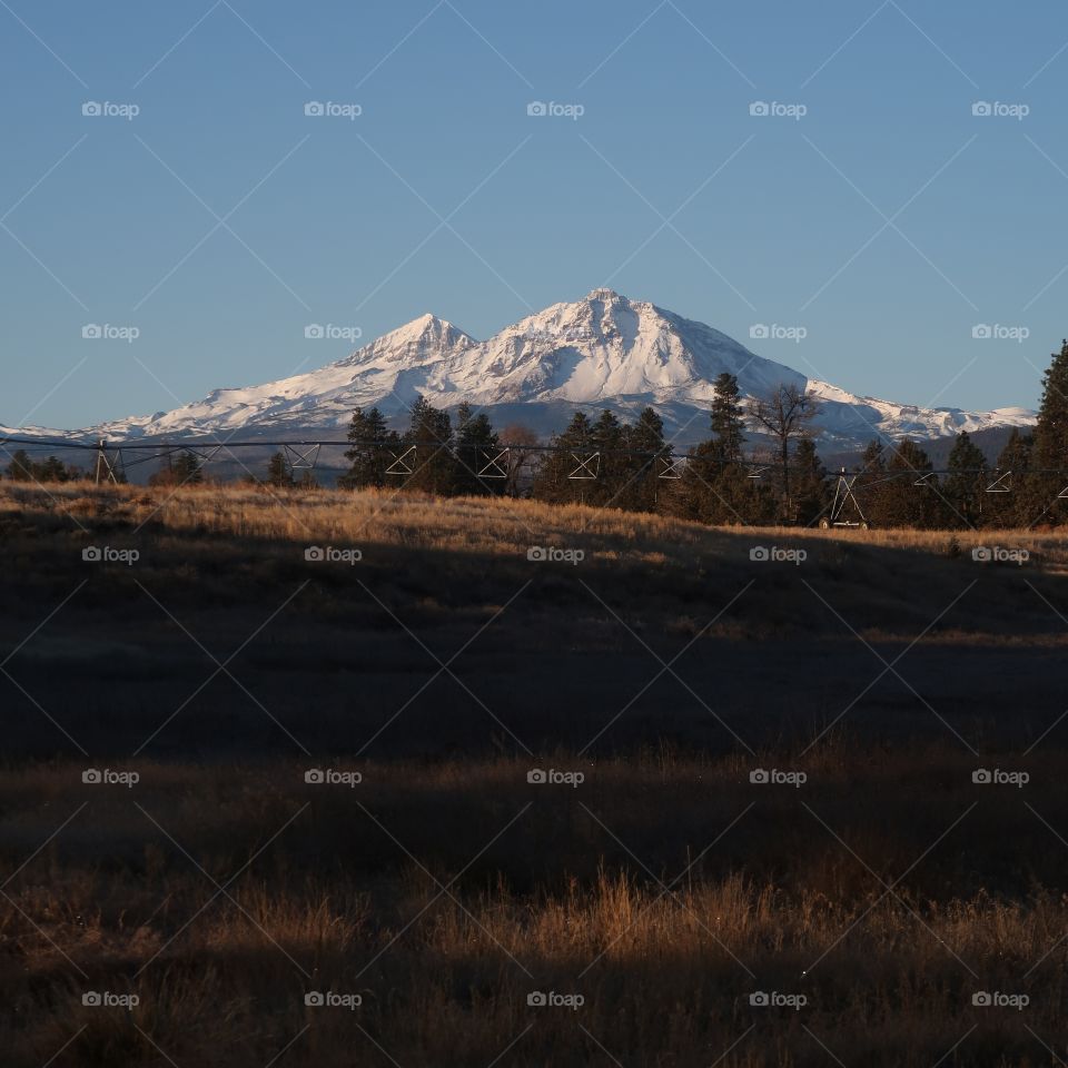 Morning light dawning on the North and Middle Sister covered in a fresh coat of snow and across a field with irrigation wheel lines in the background on a cold and clear winter morning.