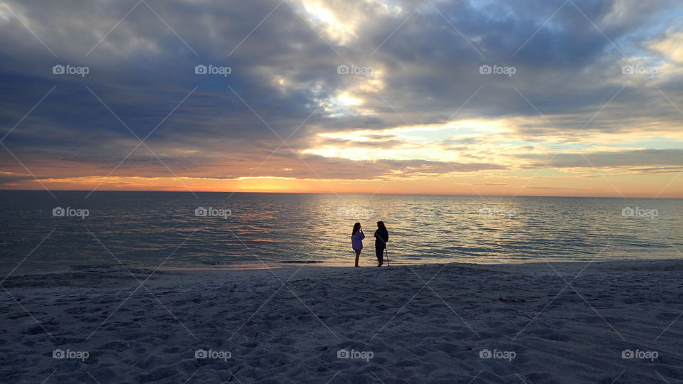 Two by seashore watching sunset speckled yellow blue gray clouds and golden horizon