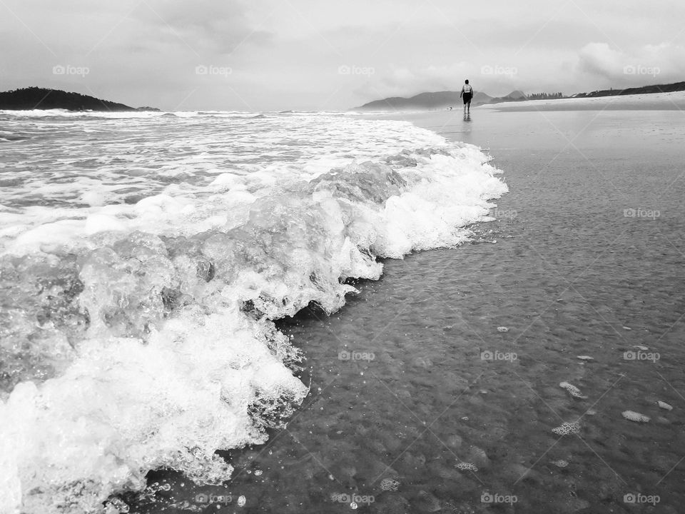 Image with angle and focus on the sea waves moving along the beach.  In the background, a lady walks on the beach, observing the natural beauty around her.