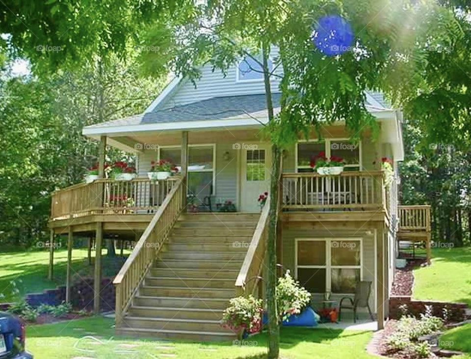 A charming two-story house is surrounded by lush greenery. The upper level features a spacious wooden deck adorned with potted flowers and railings. A wide staircase leads up to the main entrance, flanked by well-maintained landscaping. 