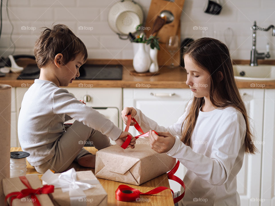 Cute toddler boy in grey pajamas smile in kitchen and packing Christmas gifts with his sister