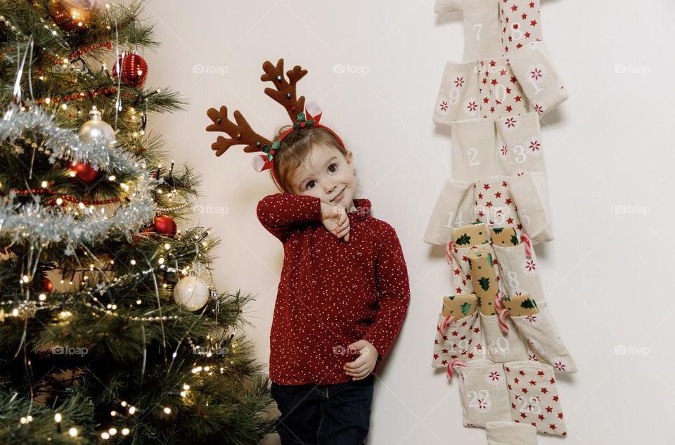 A portrait of one beautiful little Caucasian girl of two years old with a headband of deer antlers on her head stands embarrassed between an elegant Christmas tree and a wall advent calendar with gifts, close-up side view.