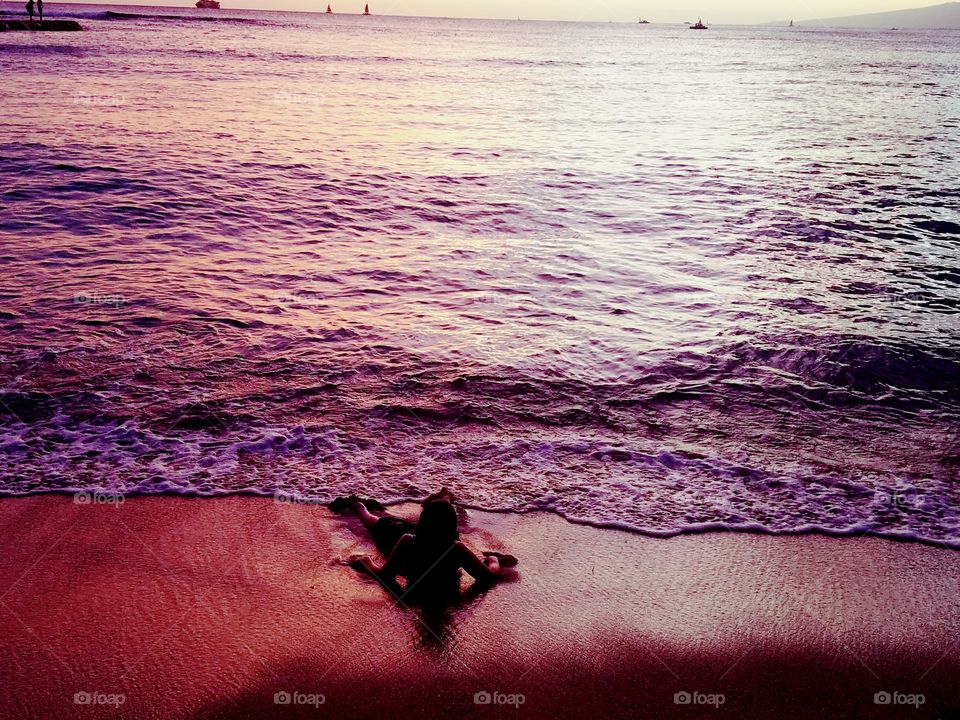 boy lays on sandy Waikiki Beach as the sun paints the ocean beautiful colors of purple pink and gold