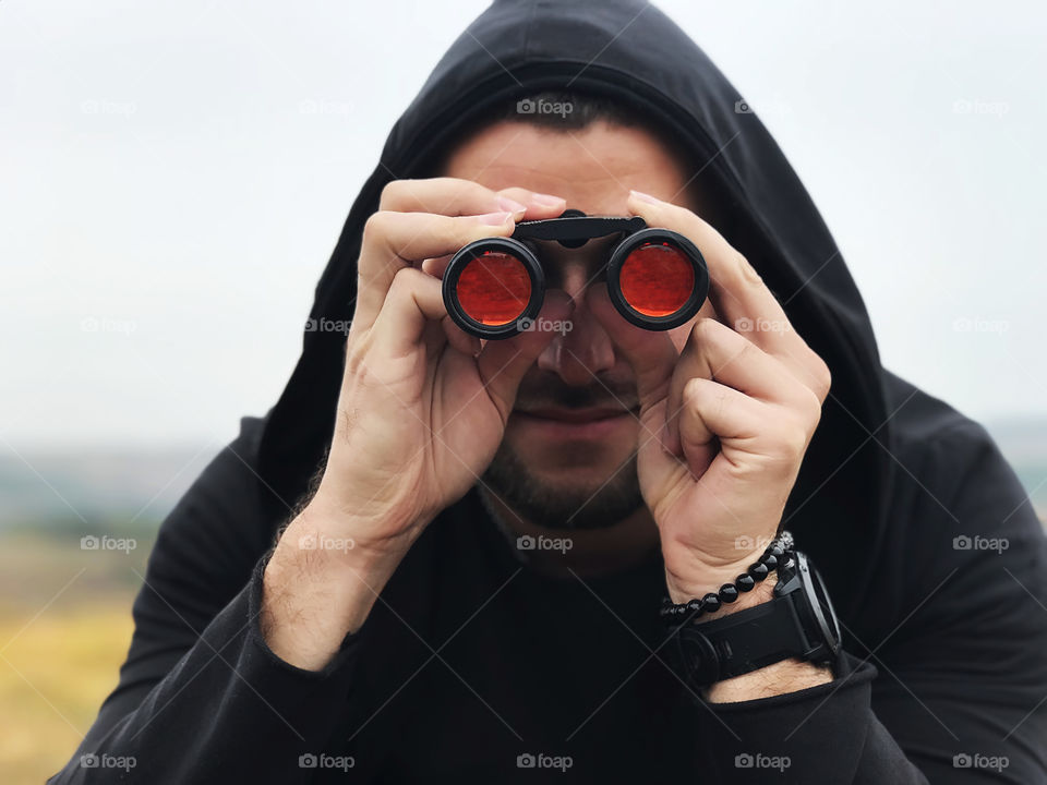 Young man wearing black hoodie, smart watches and a bracelet watching through the binoculars with red glasses 