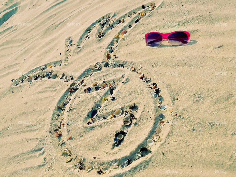 a pineapple design traced into the sand and outlined with miniature seashells with a pair of sunglasses sitting nearby
