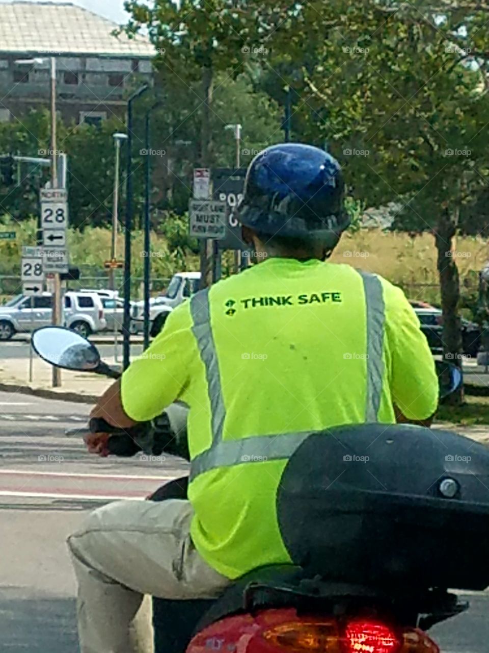 Man on scooter in city traffic with fluorescent green shirt says "think safe". Helmet black.