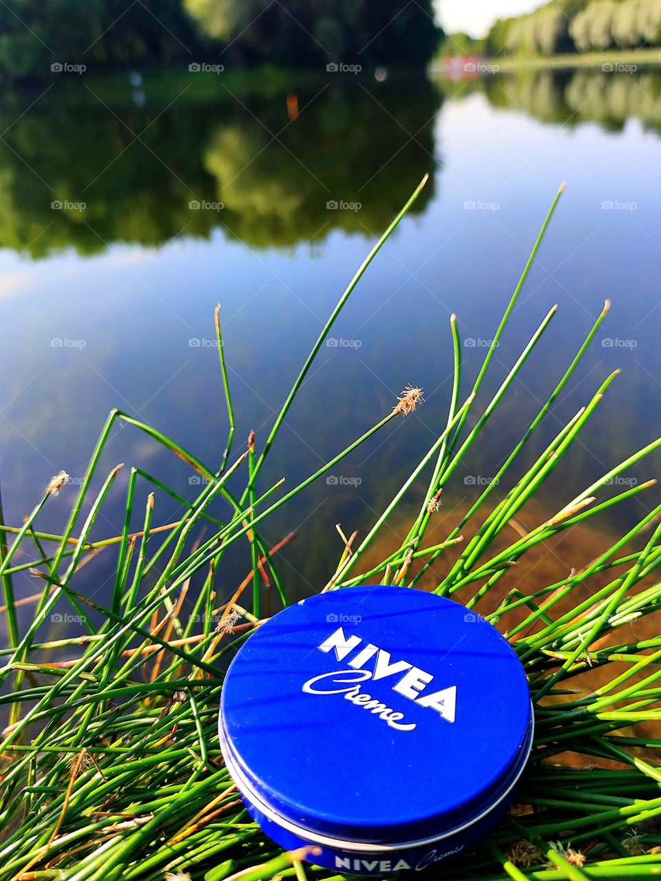 On the green grass of the river bank lies a blue box with the inscription: "Nivea cream".  In the background, trees and a blue sky with white clouds are reflected in the water.