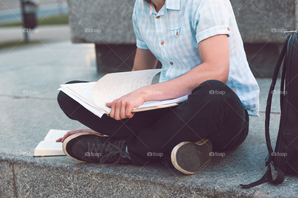 Student making the notes learning from books sitting on a monument outside of university. Young boy wearing a blue shirt and dark jeans