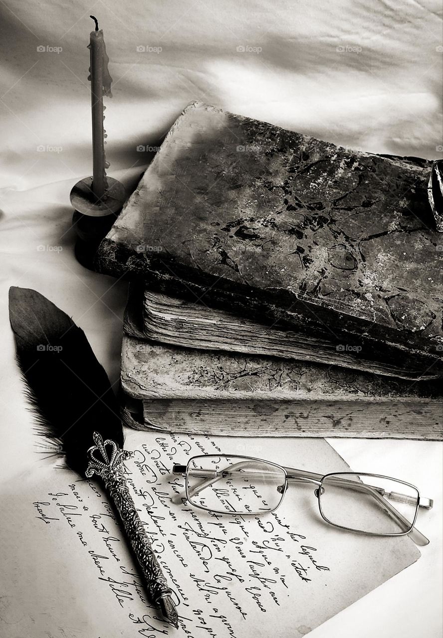 Black and white photo. Connection of generations. Old books, a piece of paper with handwritten text, a fountain pen and glasses. An extinguished candle in the background