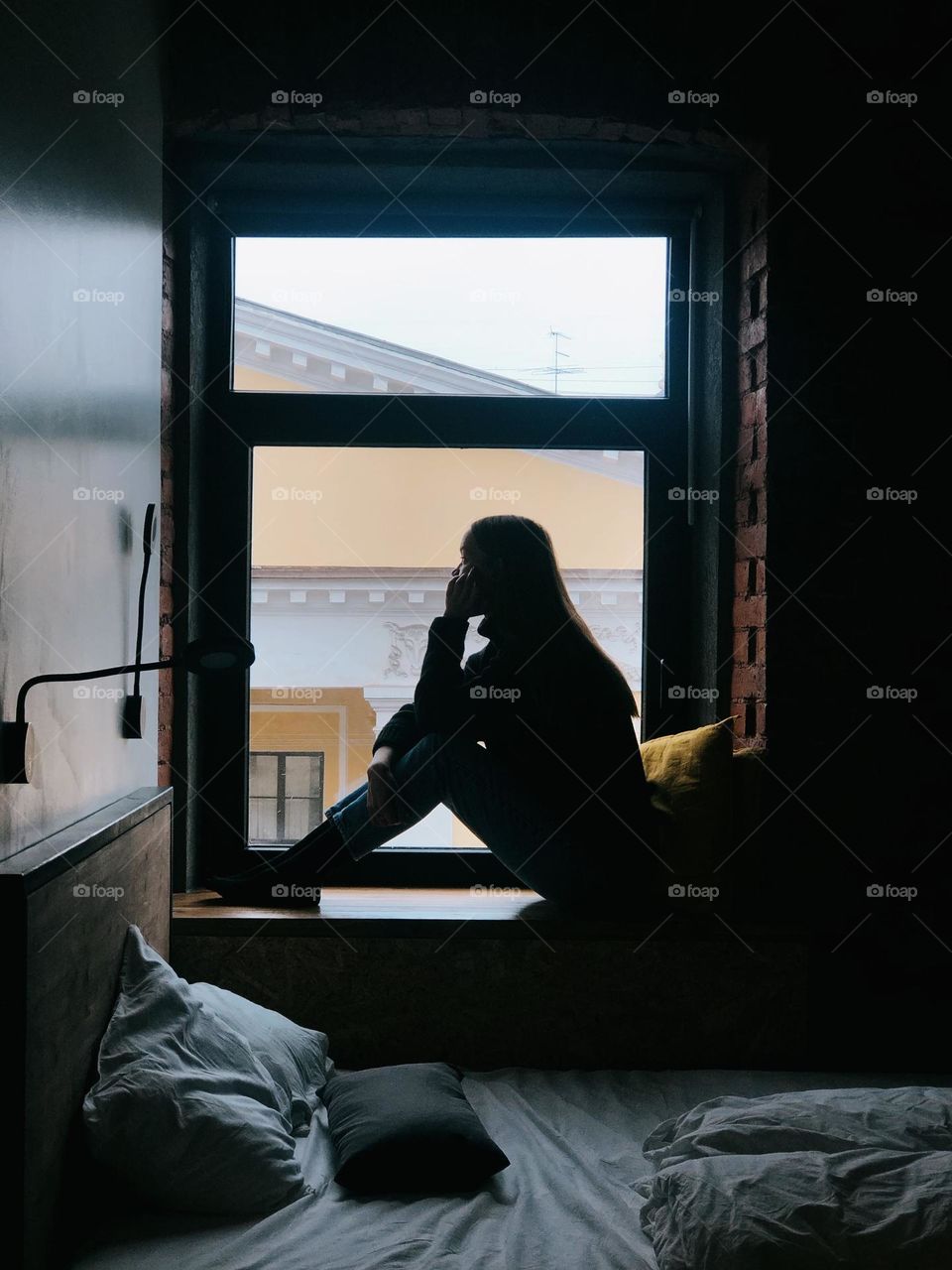 A young girl with long hair sits on the windowsill by the window and talks on the phone, the girl's silhouette