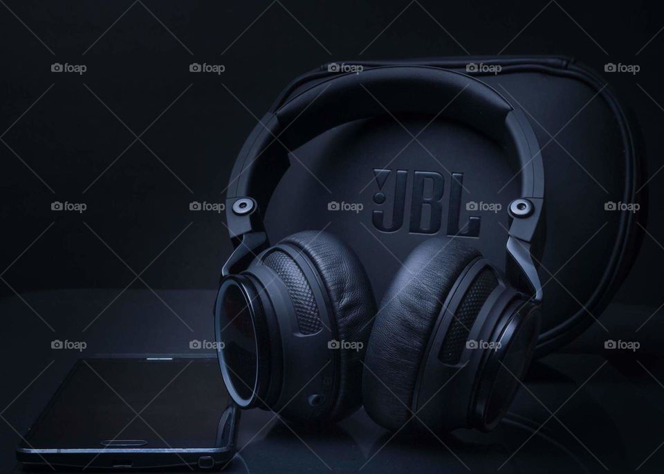 all black on black image of black headphones leaning against black headphones case next to a black cell phone on a black tabletop with a black background