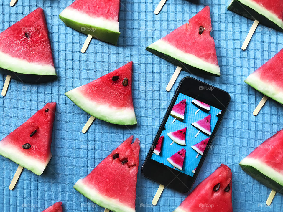 Creative colorful pattern made of slices of red watermelon triangles and mobile phone with a photo on blue textile background 