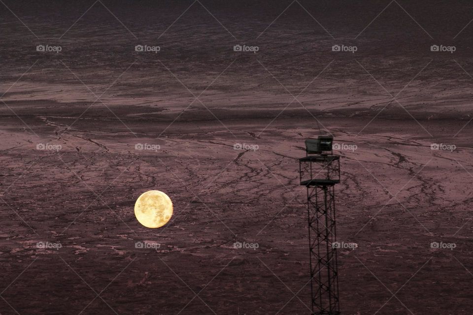 Double exposure of a picture of the moon and some tower structure with the dried up ground of a lake. It gives the picture the look of an alien planet.