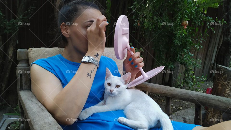 A woman is applying makeup and a small white cat is lying on her stomach