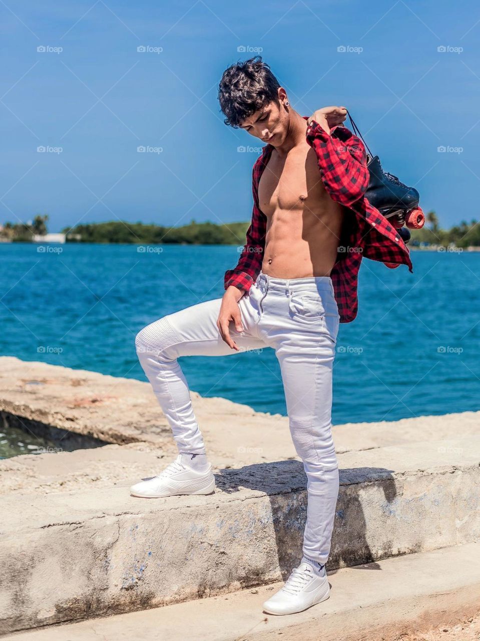 Portrait of young man standing on the beach against the background of blue water and clear sky. The person was wearing casual clothes and holding black roller skates on their shoulders