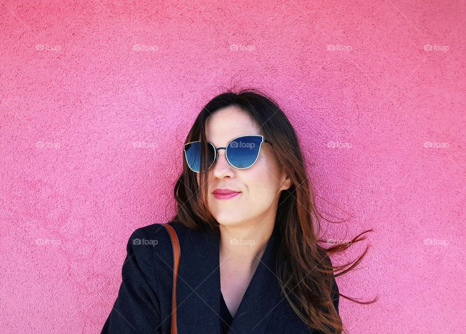 Portrait of happy young woman wearing sunglasses, standing in front of pink wall