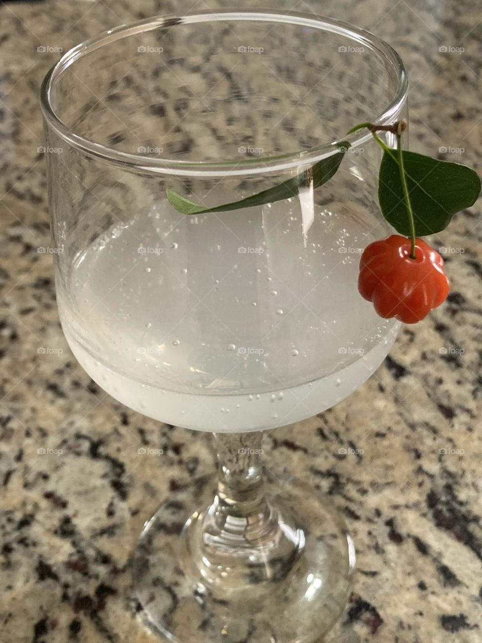 Taça com bebida branca com baixo teor alcoólico com enfeite de pitanga e folha. A bebida está sobre mesa de granito.