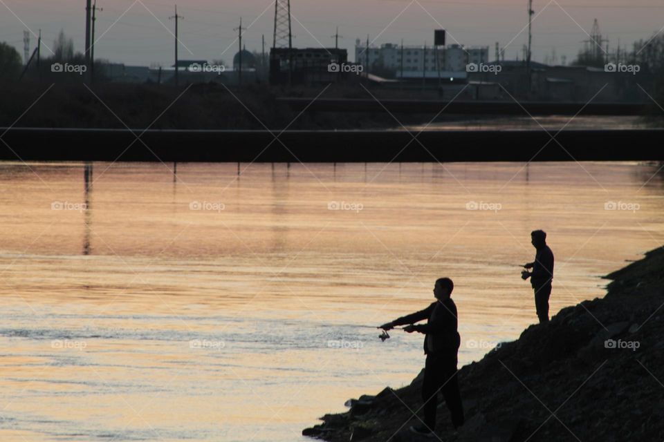 fishermen at sunset stand by the river in the background city buildings dot silhouettes.