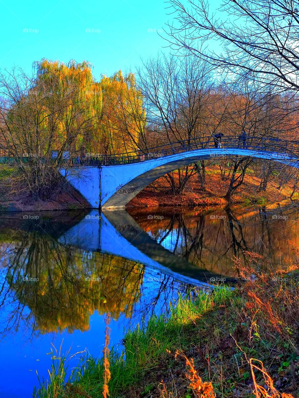 Autumn landscape. There is a stone bridge across the river, which is reflected in the blue clear water. On the shore there are autumn trees with yellow and red leaves, which are reflected in the blue water.